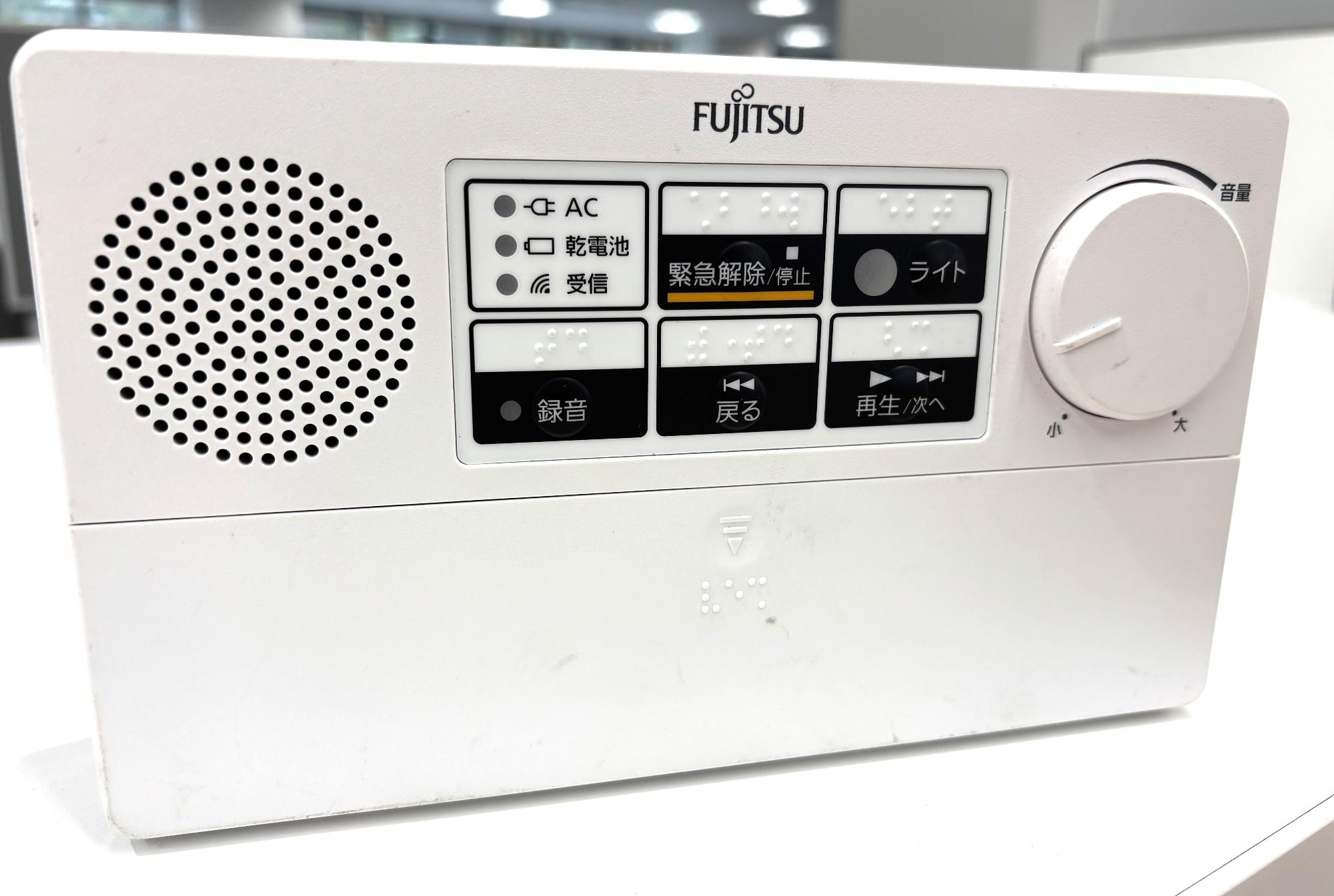 白い筐体の前面に操作パネル、スピーカーの穴、ボリュームのダイヤルがあり、上側にアンテナが伸びている富士通製の防災行政無線受信機「CR-672B型」の写真