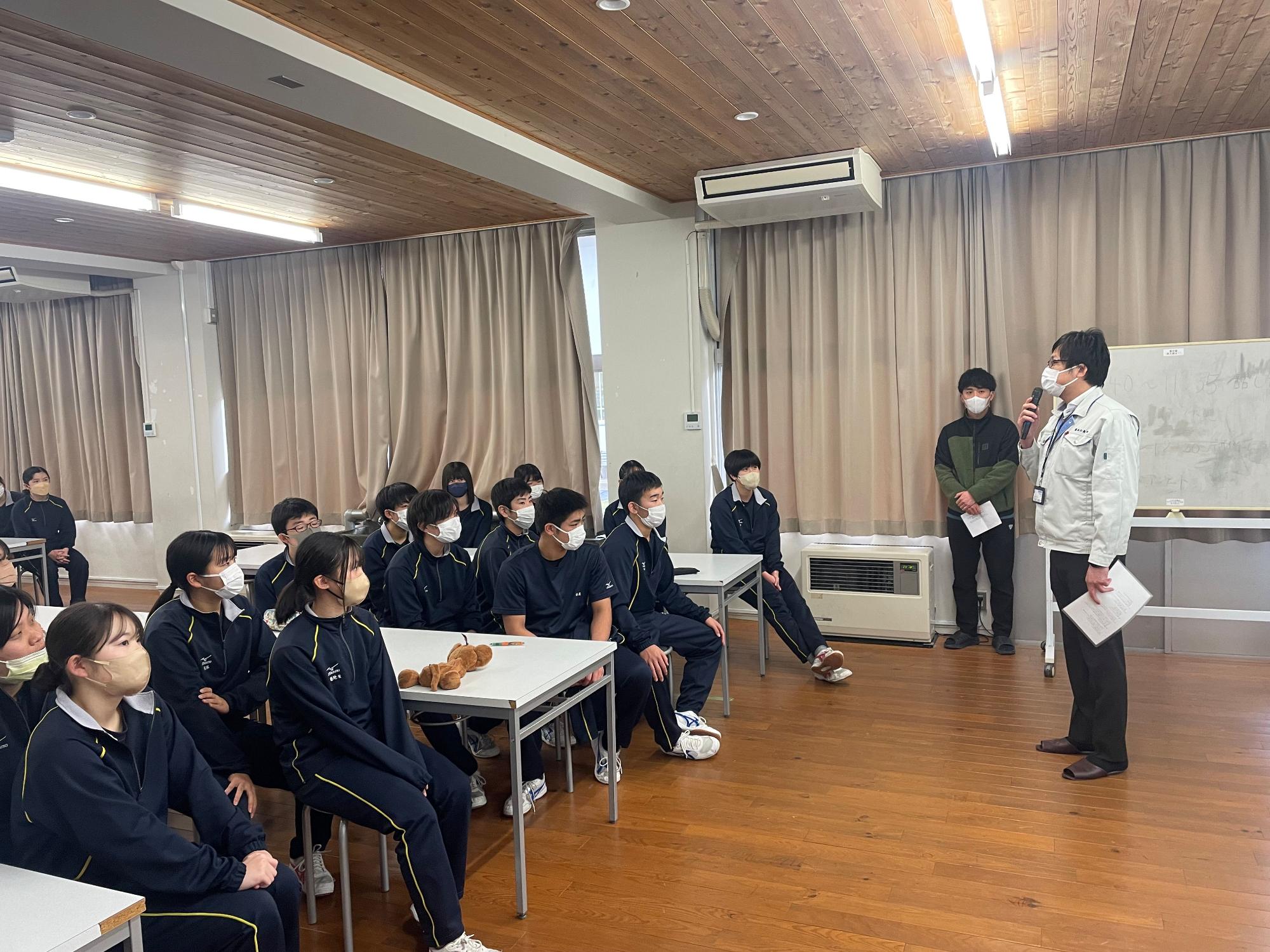 広い教室内でグループごとの机に着席し、マイクを持って立つ男性の話を聞いている生徒達の写真