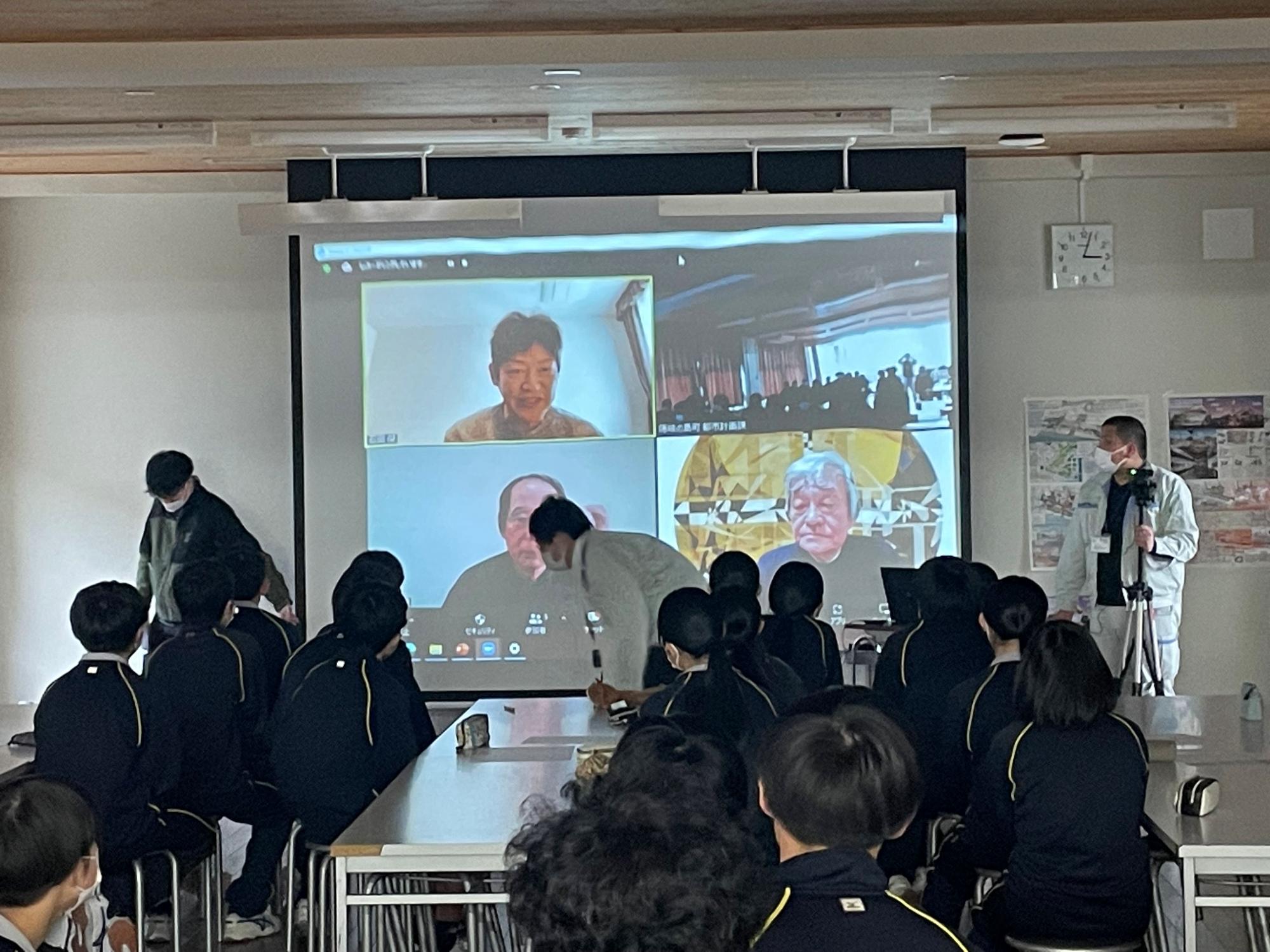 スクリーンに映し出された数名の男性達の話を、着席して聞いている生徒達の後ろ姿の写真