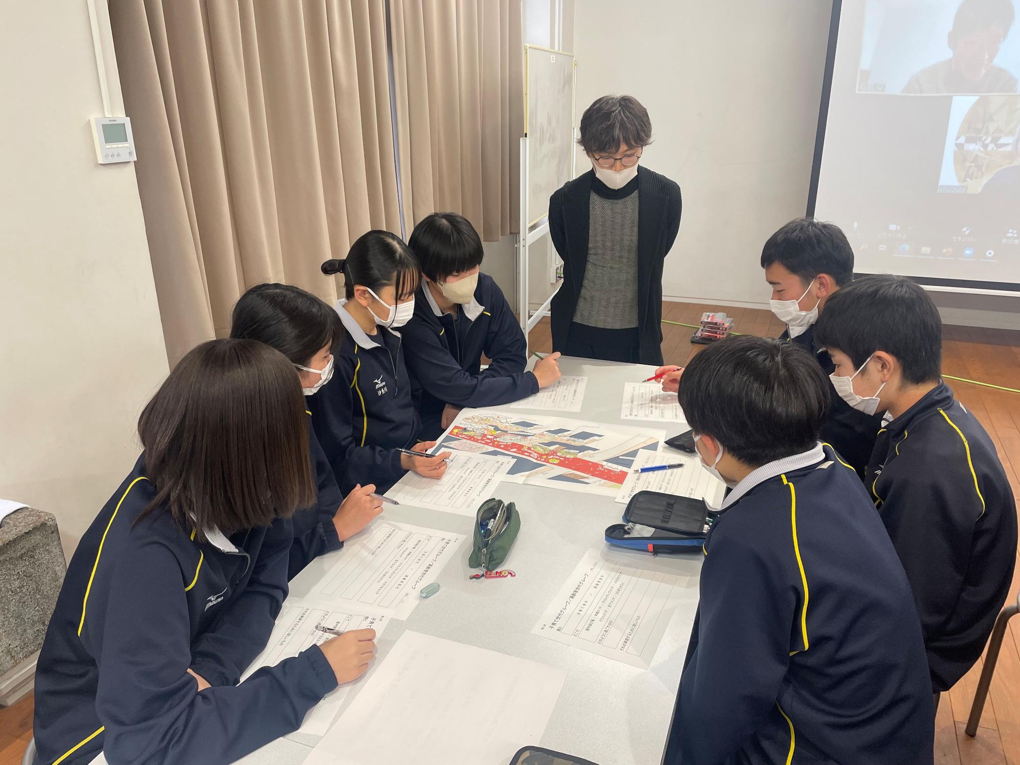 ワークシートなどを上に広げた長机を囲み話し合いをする生徒達と、側に立って見守る女性の写真