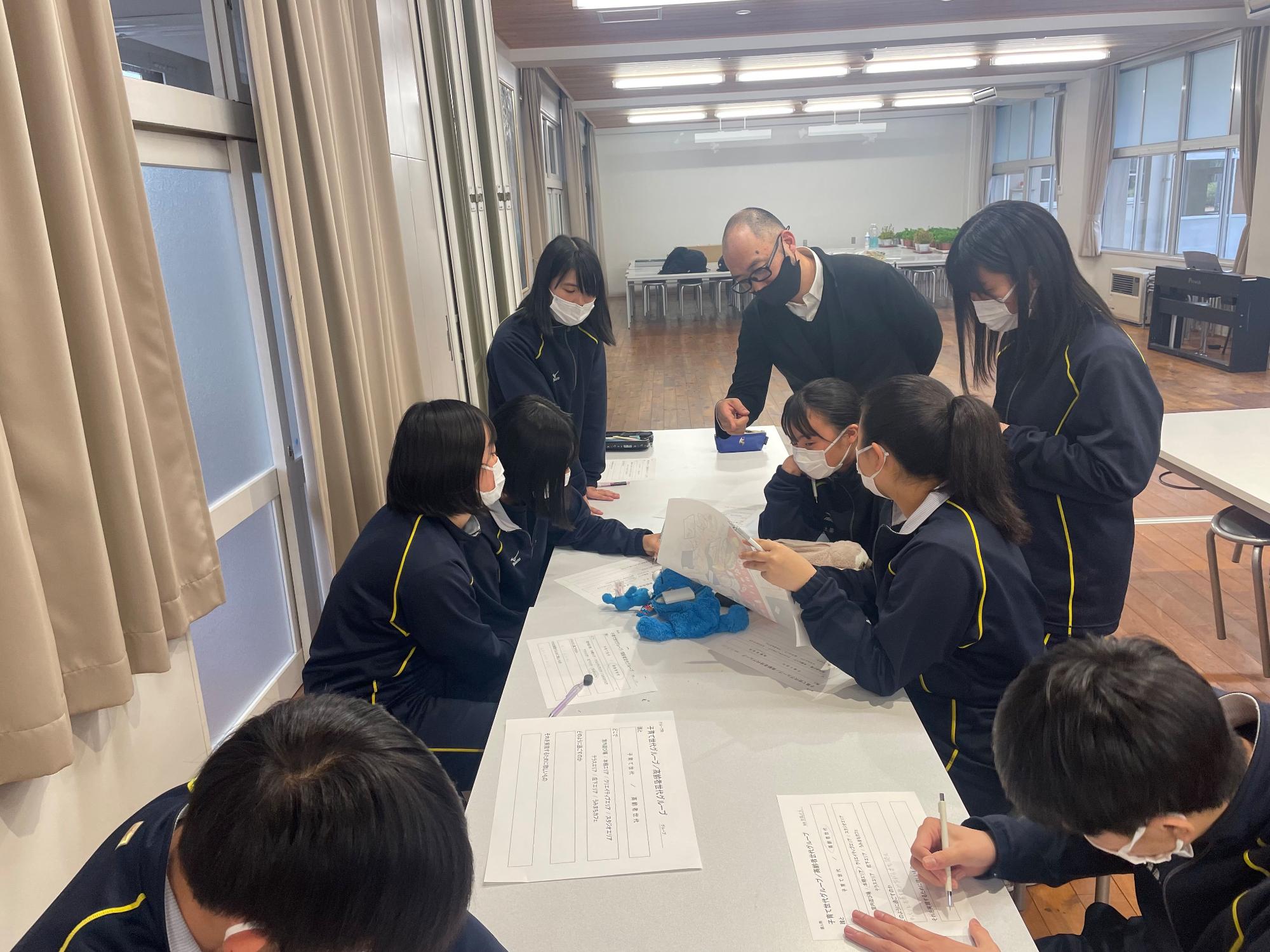 長机を囲んで集まり、話し合いやワークシートへ書き込みを行う生徒達の写真