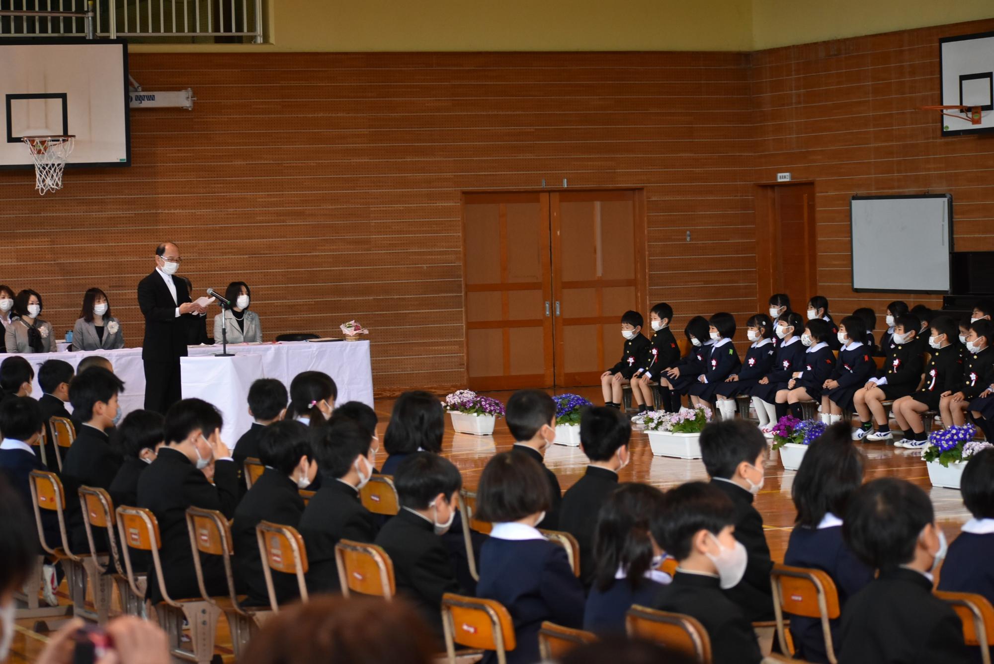 体育館で校長先生が座っている新入生に向けて話をしている写真