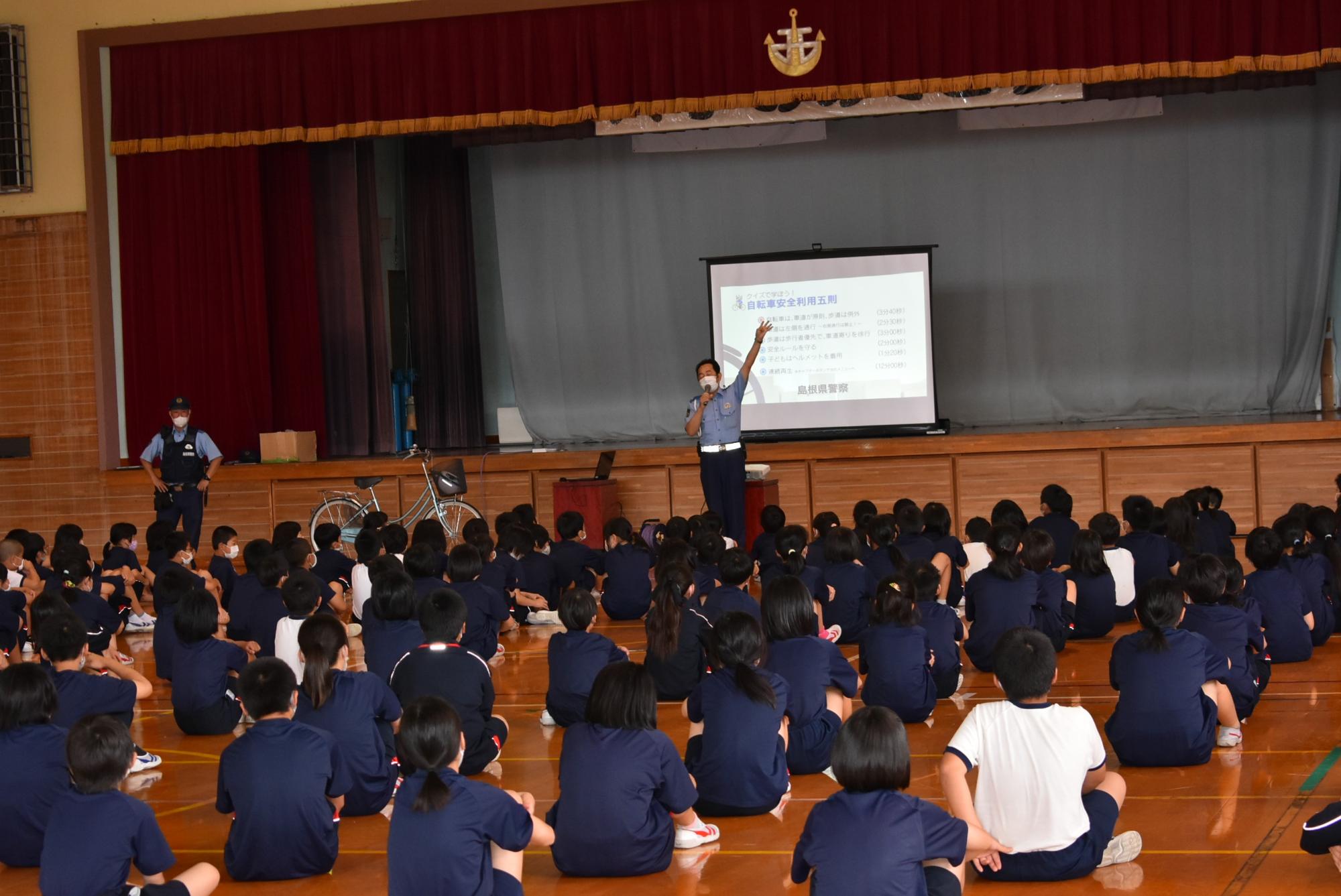 体育館で警察官が座っている児童たちに交通安全教室を行っている写真