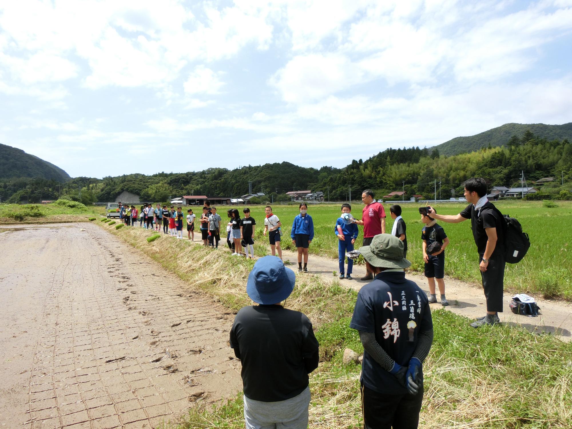 田んぼを前に横一列に並び、先生の指導に耳を傾けながら立っている児童たちの写真