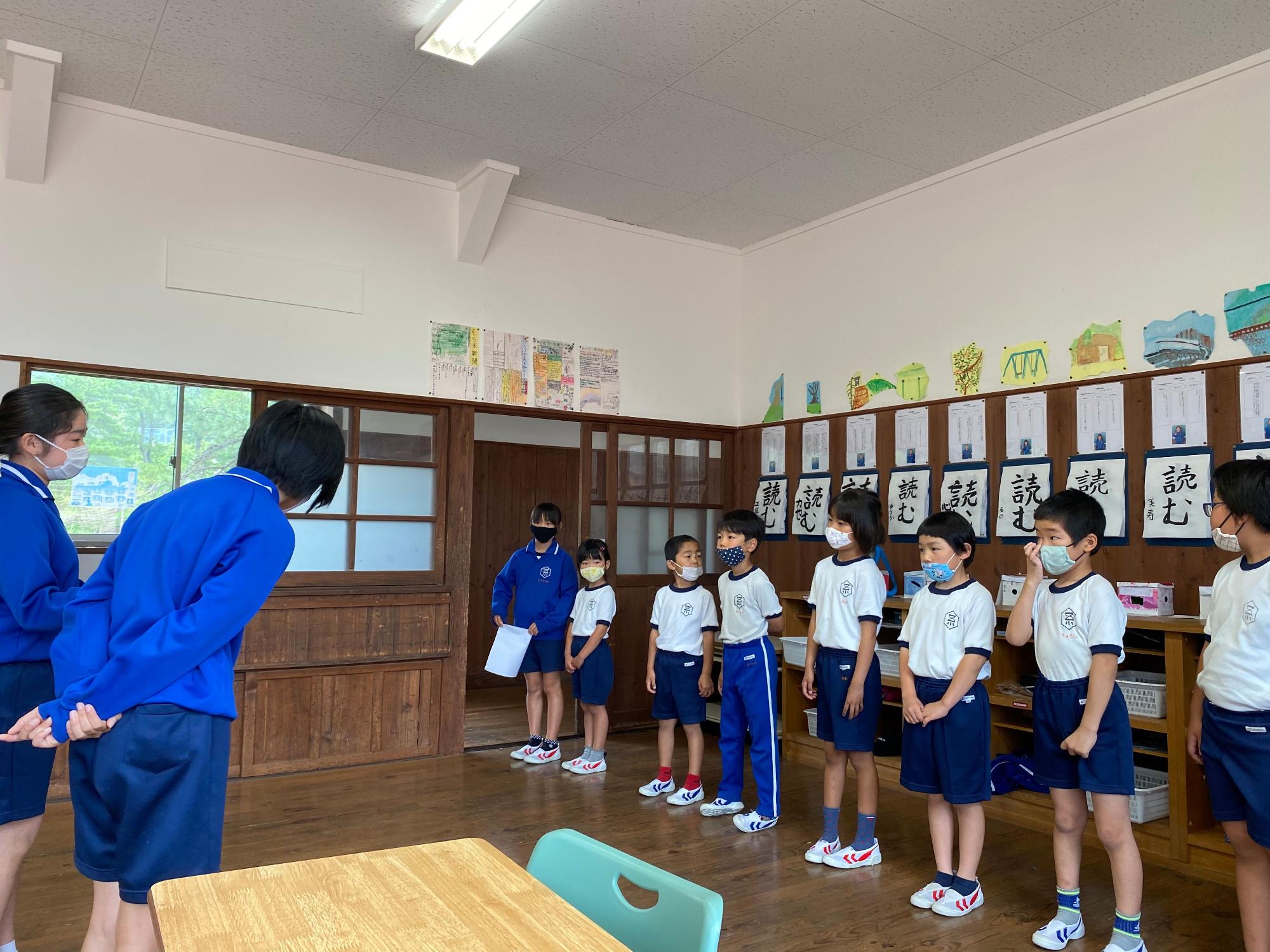 教室で体操着姿の児童二人が前に立ち、うち一人が向かい合って横に並ぶ児童たちに挨拶をしている様子の写真