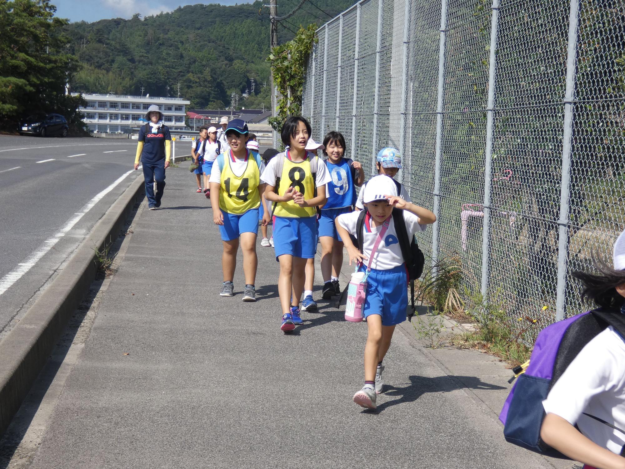 大きな荷物に水筒を持って道路を元気に歩く生徒たちの遠足の写真
