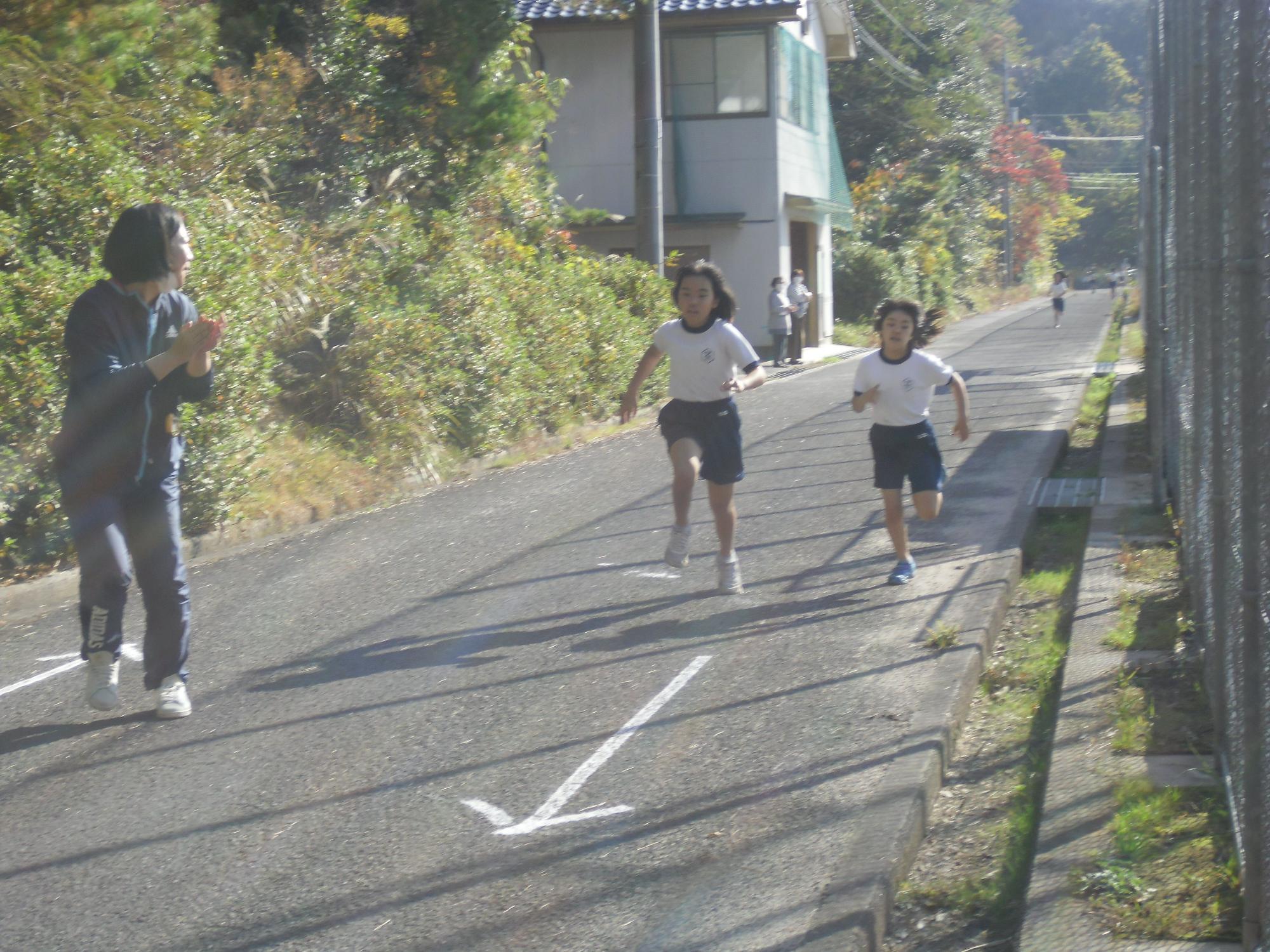 一人に女性が手を叩いて応援する中、道路を体操着姿の児童二人が一生懸命走っている写真