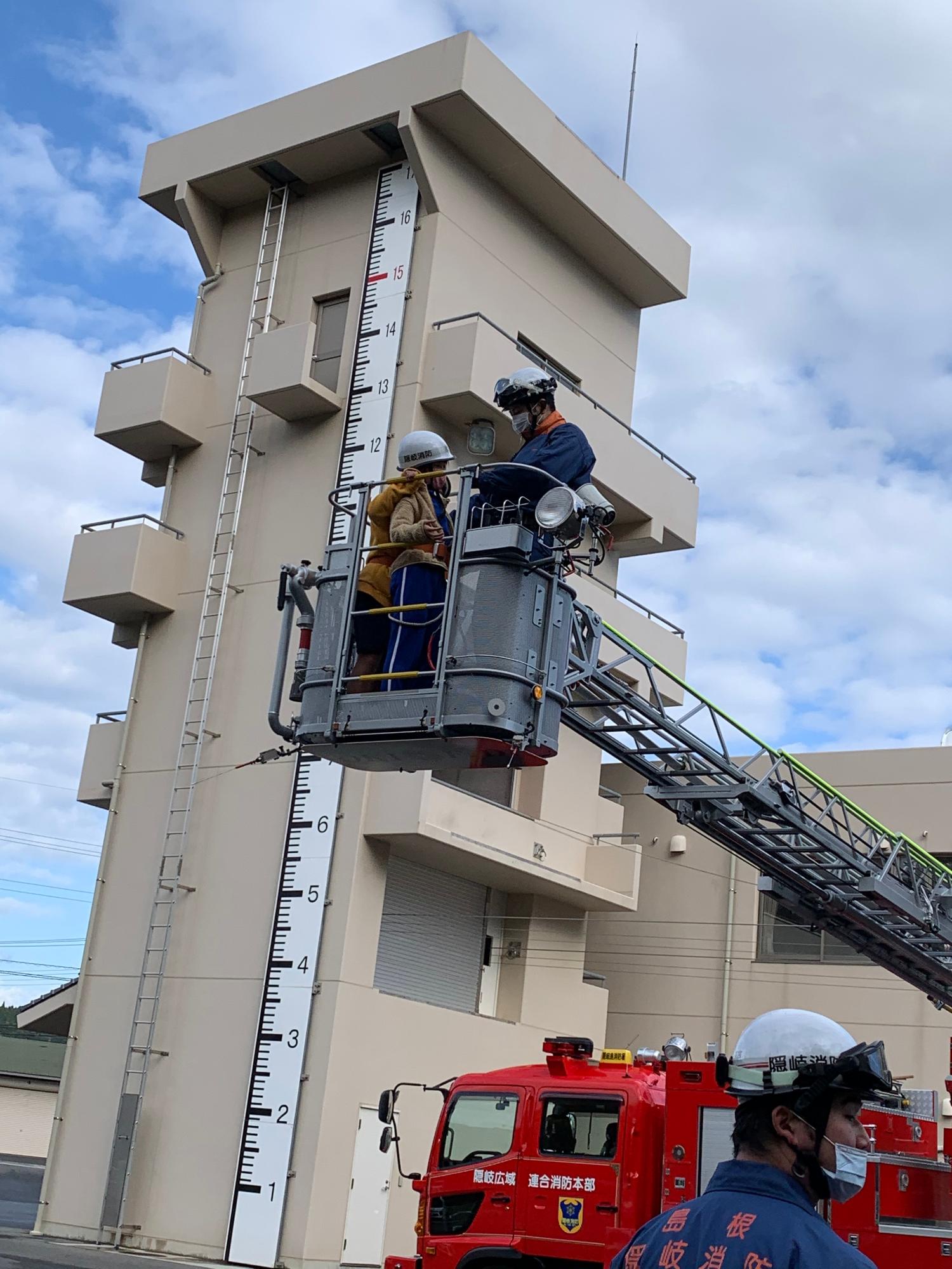 大きな塔の脇に止めたはしご車のかごに消防士の方と一緒に乗っている様子の写真