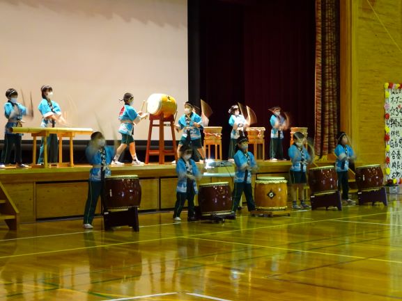 体育館のステージで太鼓がいくつも並べられ、法被姿の生徒たちがそれぞれ太鼓を叩いている写真