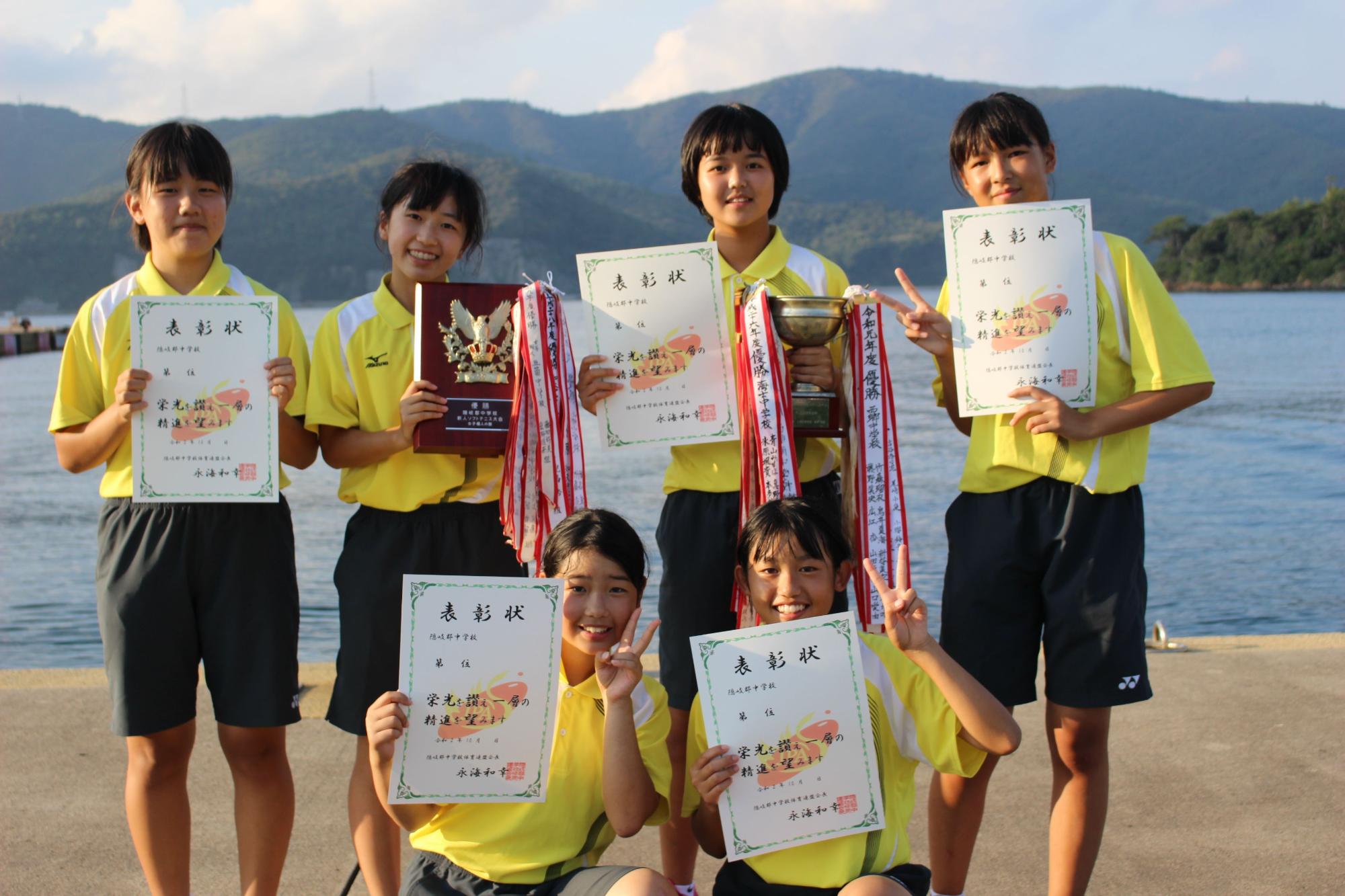 海辺で女子生徒たちが賞状とトロフィーを持って笑顔で写る写真