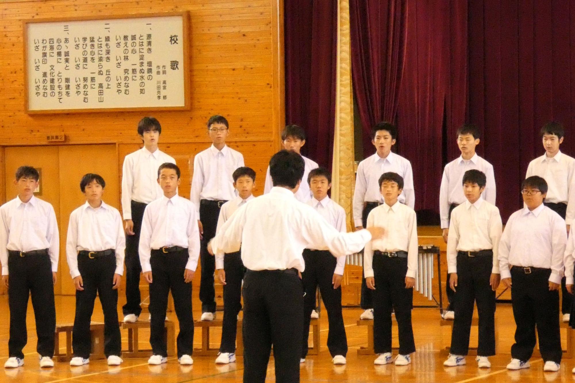 体育館で男子生徒たちが指揮者に合わせて合唱している写真