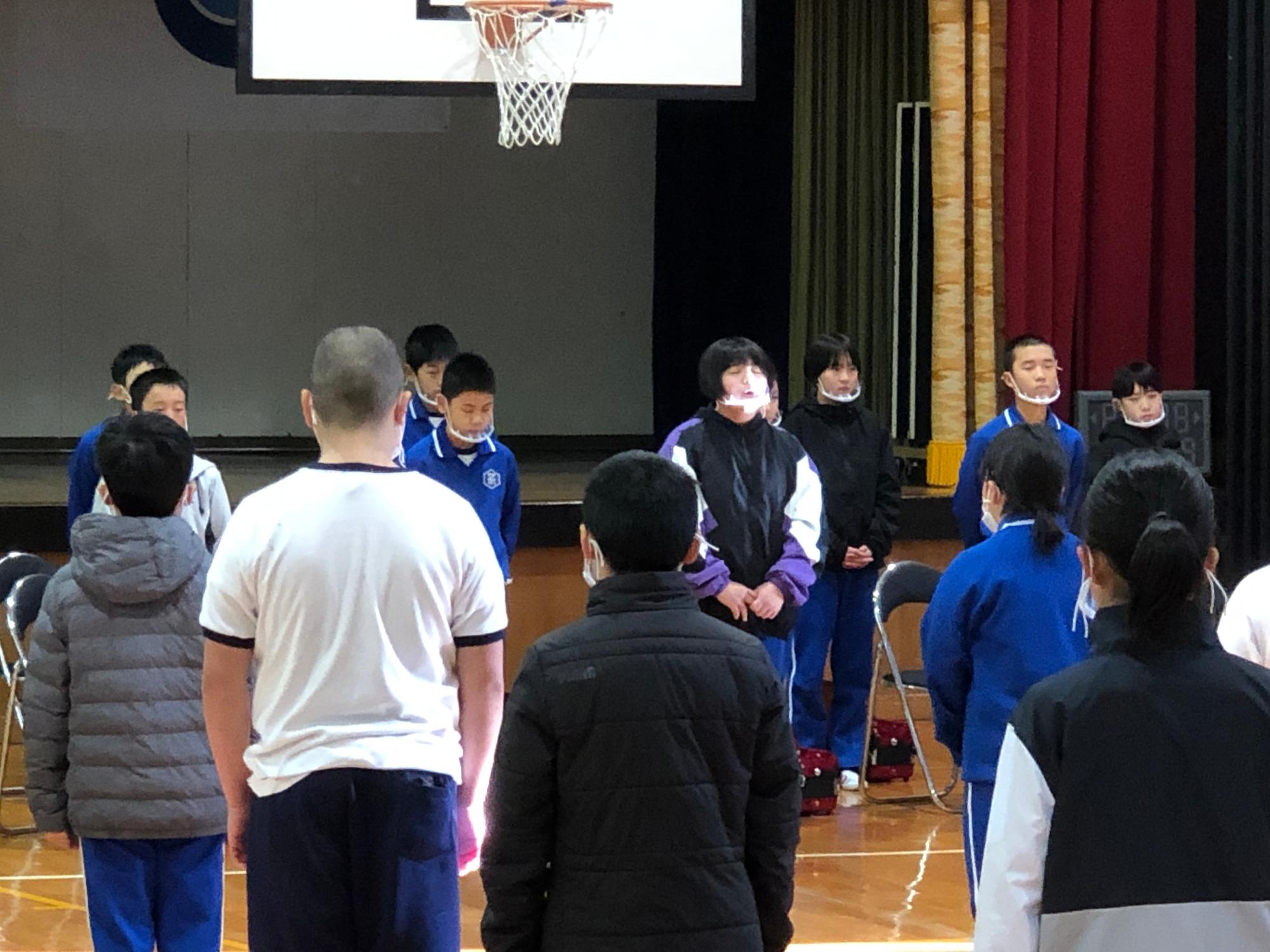 体育館で児童たちが起立をして引き継ぎ式を行っている写真