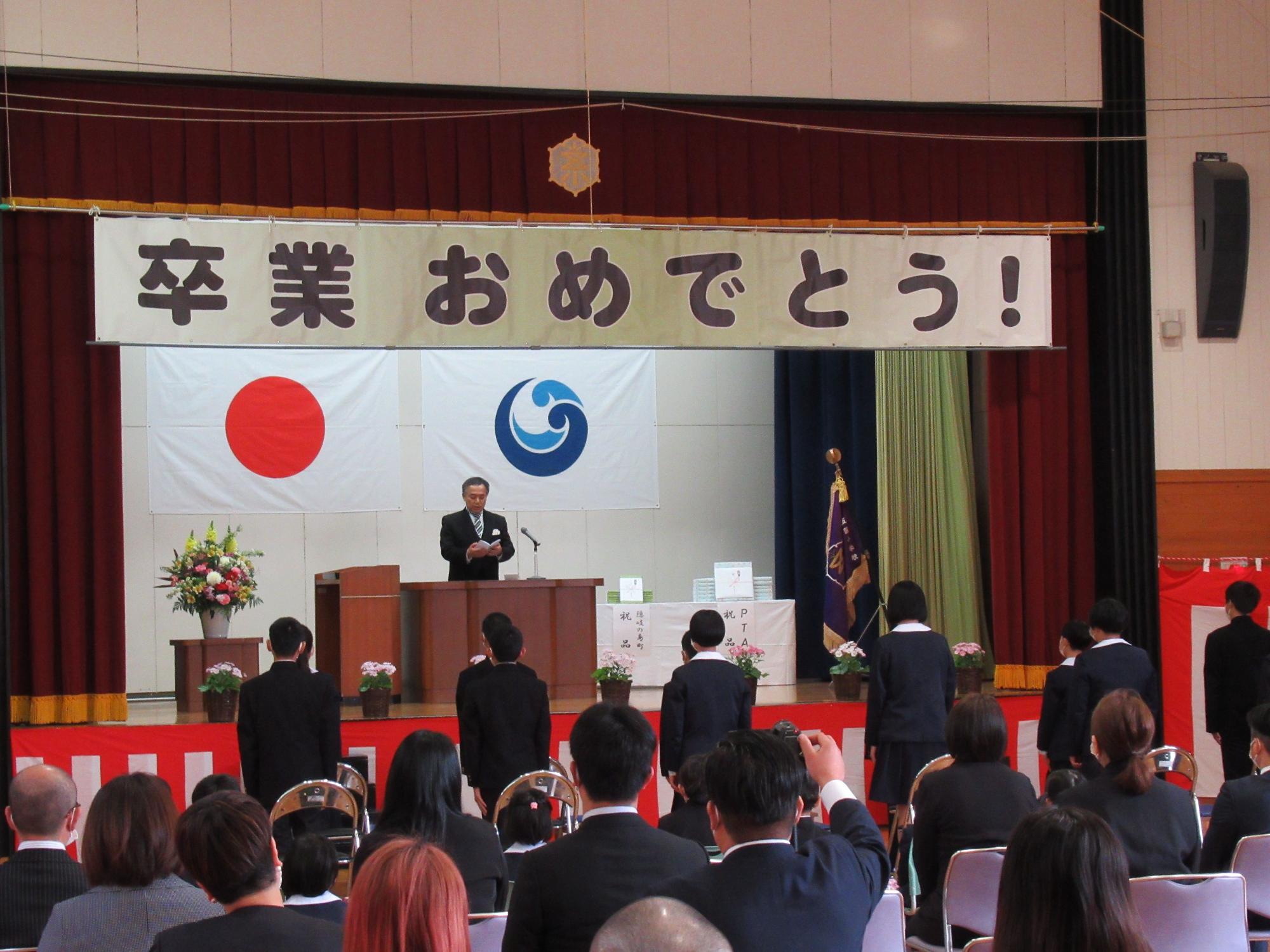 卒業証書授与式でステージの上に立つ教職員が児童たちに対して書類を見て読んでいる様子の写真