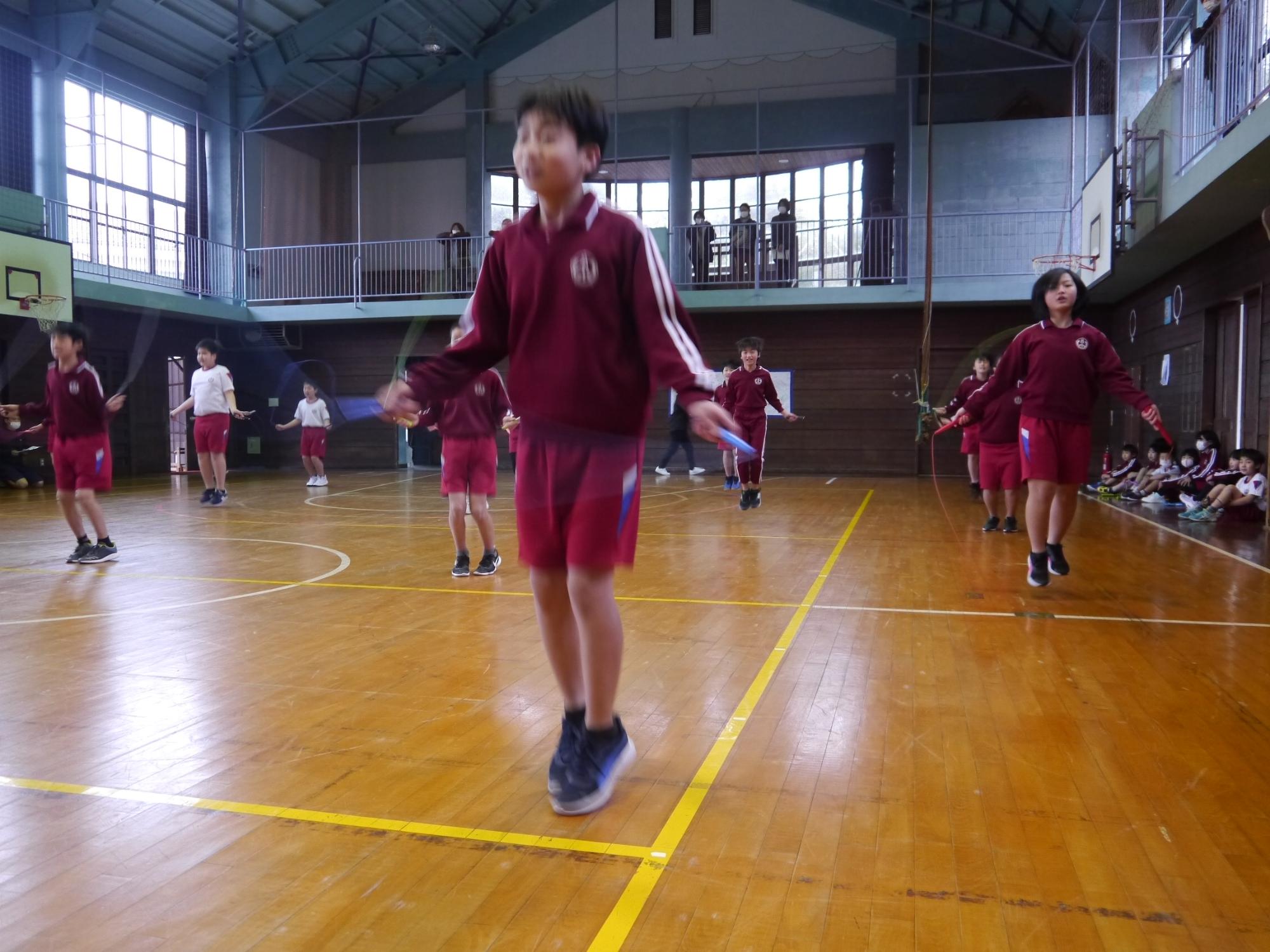 体育館で赤いジャージを着た児童たちがなわとびをしている写真
