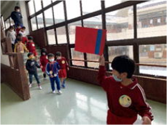 廊下で児童が赤に青い線が入った旗を掲げて先頭を歩き、後ろに列が続いている写真