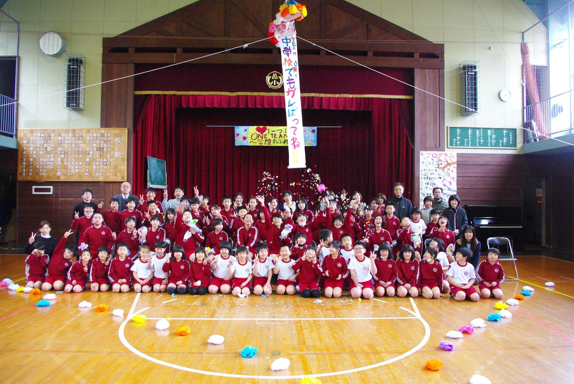 体育館の舞台前で全校の児童と教職員がそろって記念撮影をしている写真