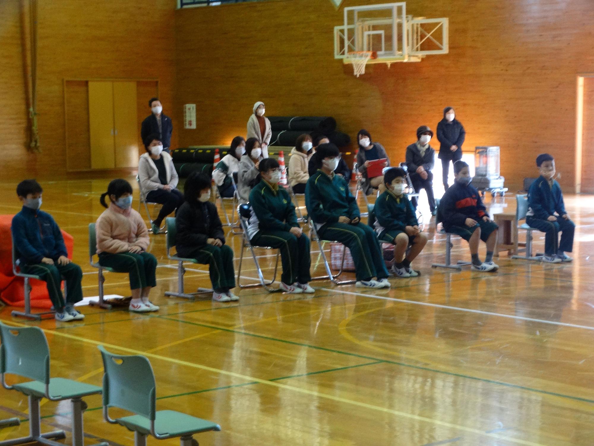 体育館で児童が横一列に並んでパイプ椅子に座っている写真