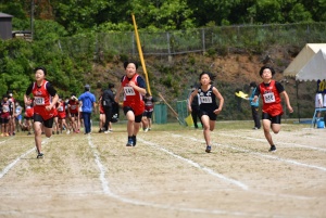 運動場で赤や黒のユニフォームを着た男子児童4人が走っている写真