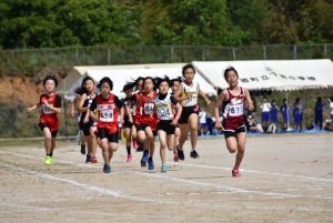 運動場で赤、黒、白のユニフォームを着た児童たちが競い合って走っている様子の写真