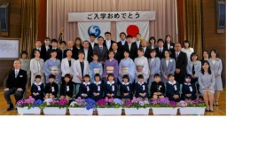 体育館の舞台前で新入生と保護者、教職員が並んで記念撮影をしている写真