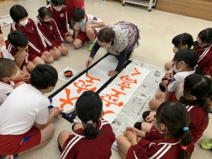 教室で先生と一緒に大きな紙に文字を書いている児童たちの写真