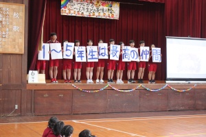 体育館の舞台上で「1〜5年生は最高の仲間」と書かれた横断幕を持つ児童たちの写真