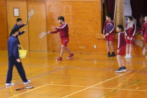 体育館で赤いジャージを着た生徒たちがバドミントンの練習をしている写真