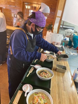 エプロンを着て、机に並べられたラーメンの盛りつけを行う生徒の写真