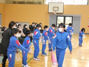 体育館で横一列になって並んでいる児童たちが、歩く児童に何かを渡している様子の写真