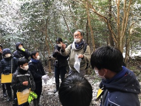 雪が残る森の中で先生の話を聞くマスクをした児童たちの写真
