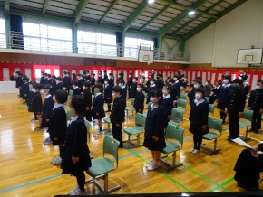 体育館でマスクをした制服姿の児童たちが椅子から一斉に起立している写真