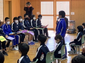 横一列に並んで座る6年生に向けて、5年生の男子児童が挨拶をしている様子の写真