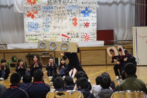 体育館の舞台の上に吊るし張られたメッセージを記した大きな紙と、その前で手作りの仮装を身につけて出し物を演じている児童たちの写真