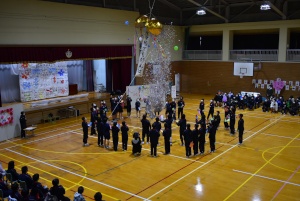 体育館の高い天井から吊された金色のくす玉が割られて開き紙吹雪が舞う中で、それを輪になって囲んで見上げ眺める児童たちの写真