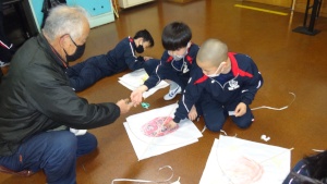 屈み込んで目線を揃えて凧作りを教えている男性と、見守られながら床に広げた凧に絵を描き込んでいる児童たちの様子の写真