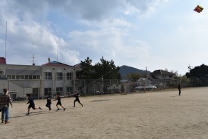 よく晴れた空の下の校庭で空に浮き上がる凧を引きながら走る児童たちの凧揚げの様子の写真