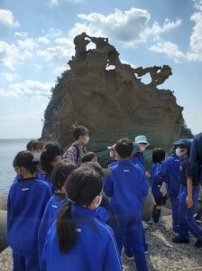 海辺の奇妙な形をした岩を前に説明を聞く子どもたちの写真