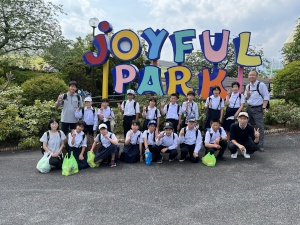 遊園地の入口でジョイフルパークと英語で書かれた看板前に並ぶ児童たちの写真