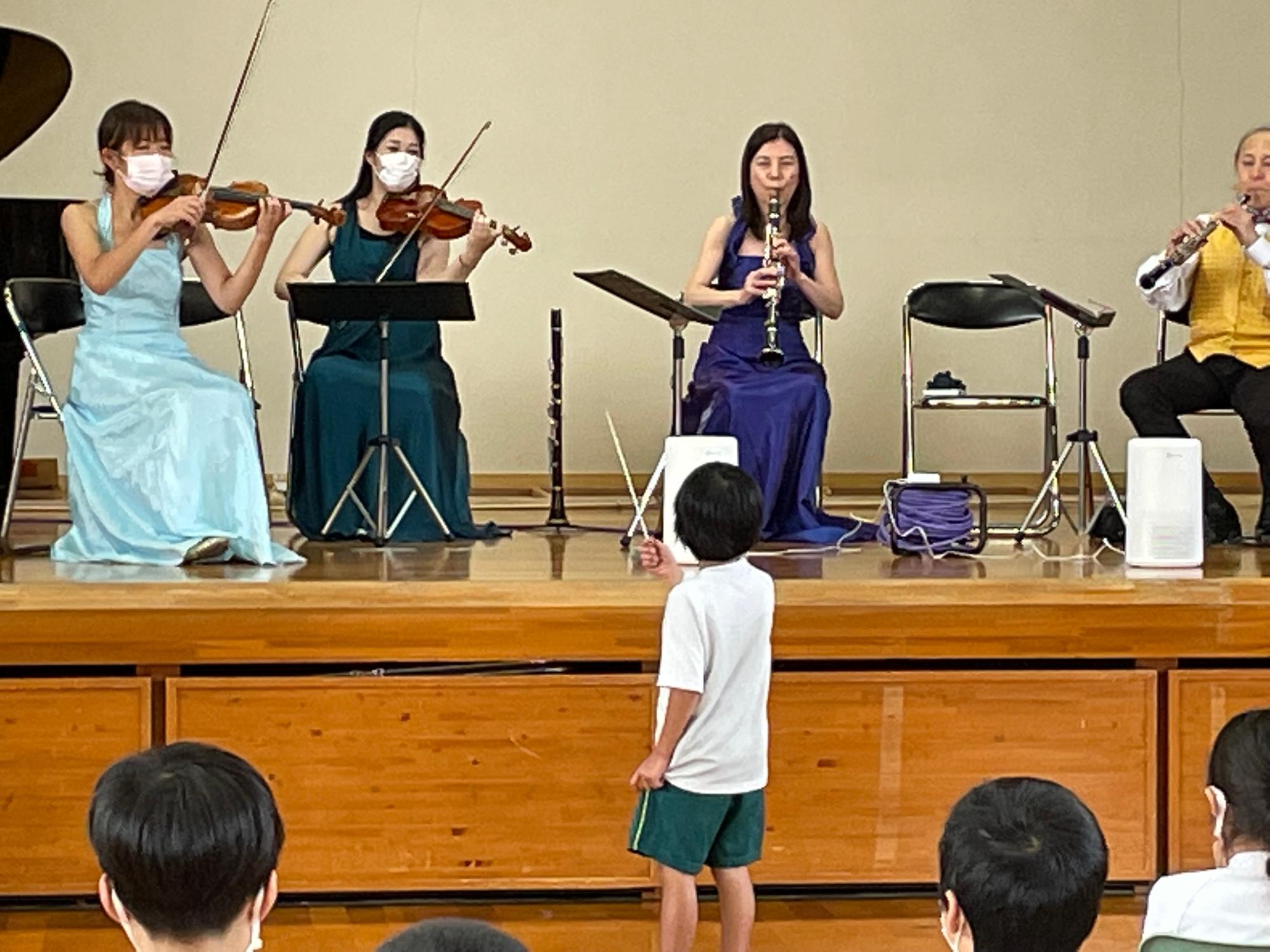 ドレスを着て演奏しているプロの音楽家の前に立ち指揮をする男子児童の写真