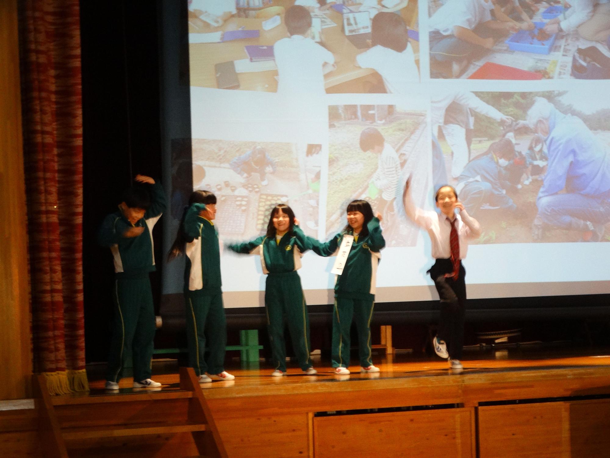 大きなスクリーンに映像が映し出されているステージの上で出し物をする児童たちの写真