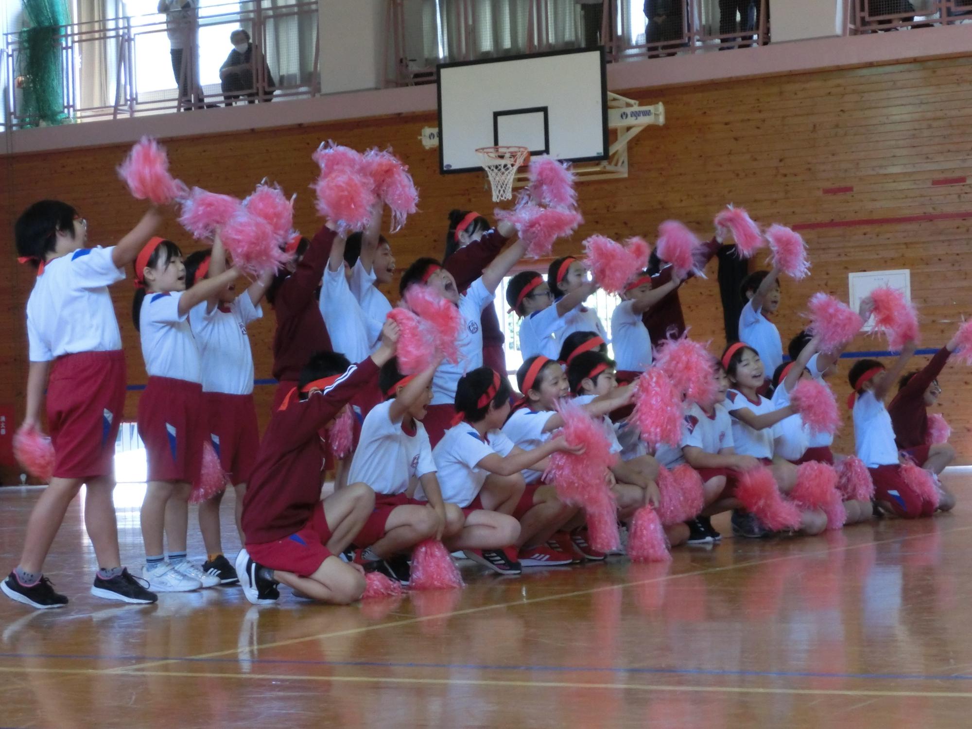 体育館で児童たちがピンクのポンポンを持って応援している写真