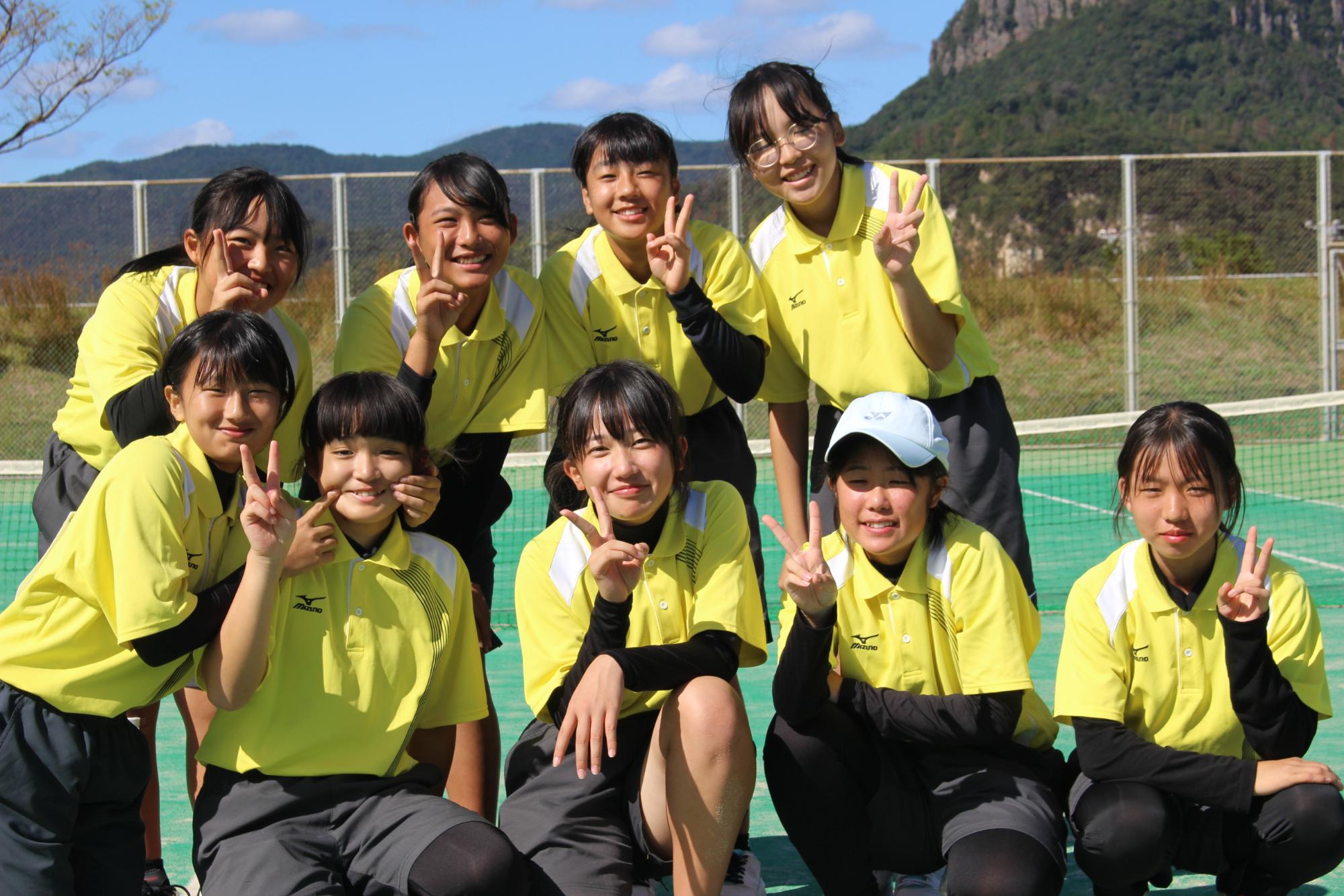 テニスコートで黄色のユニフォームを着た女子生徒が2列に並んでピースをしている写真