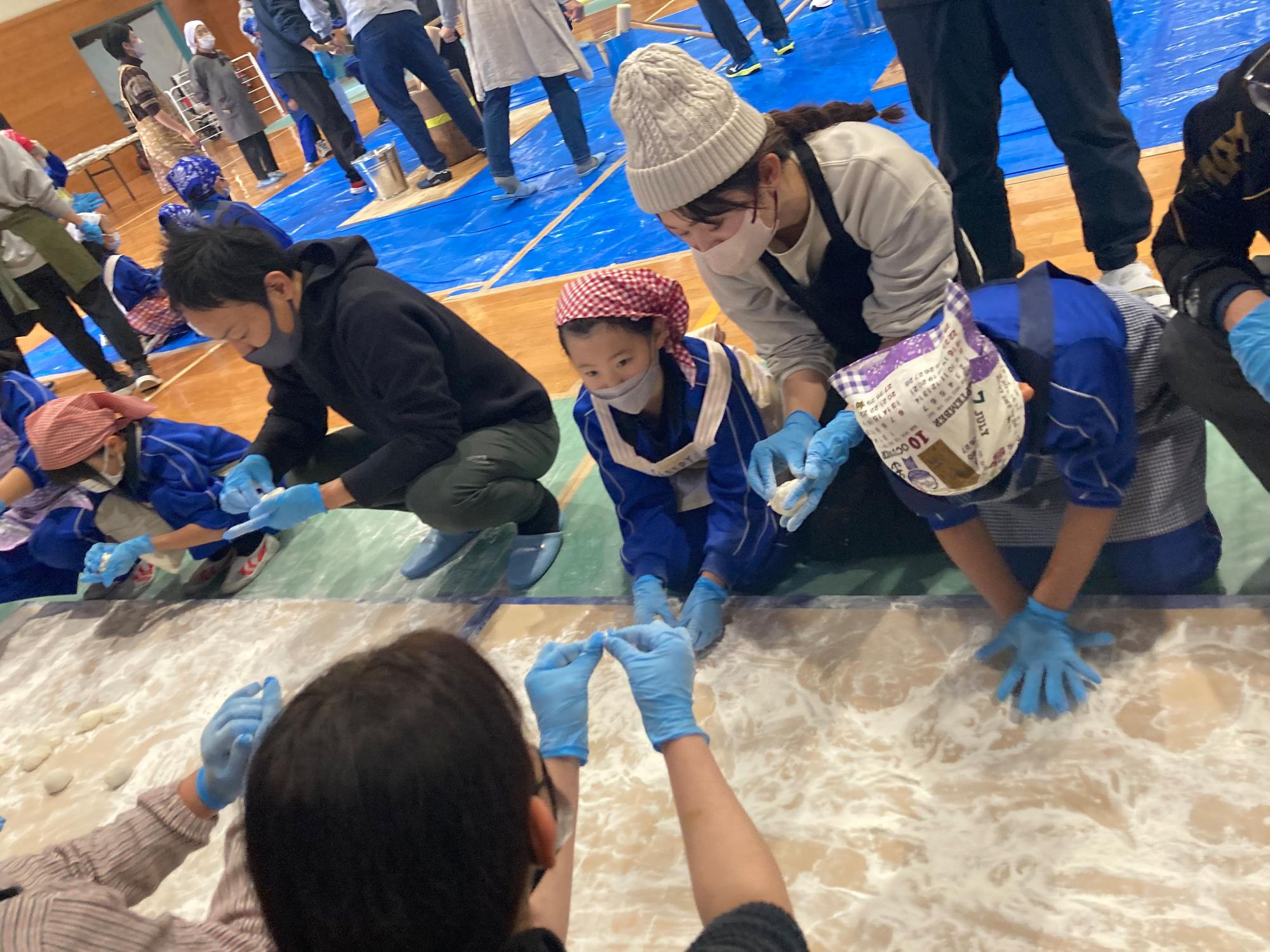 体育館で大人と児童が一緒に餅を摘まんで形を整えている写真