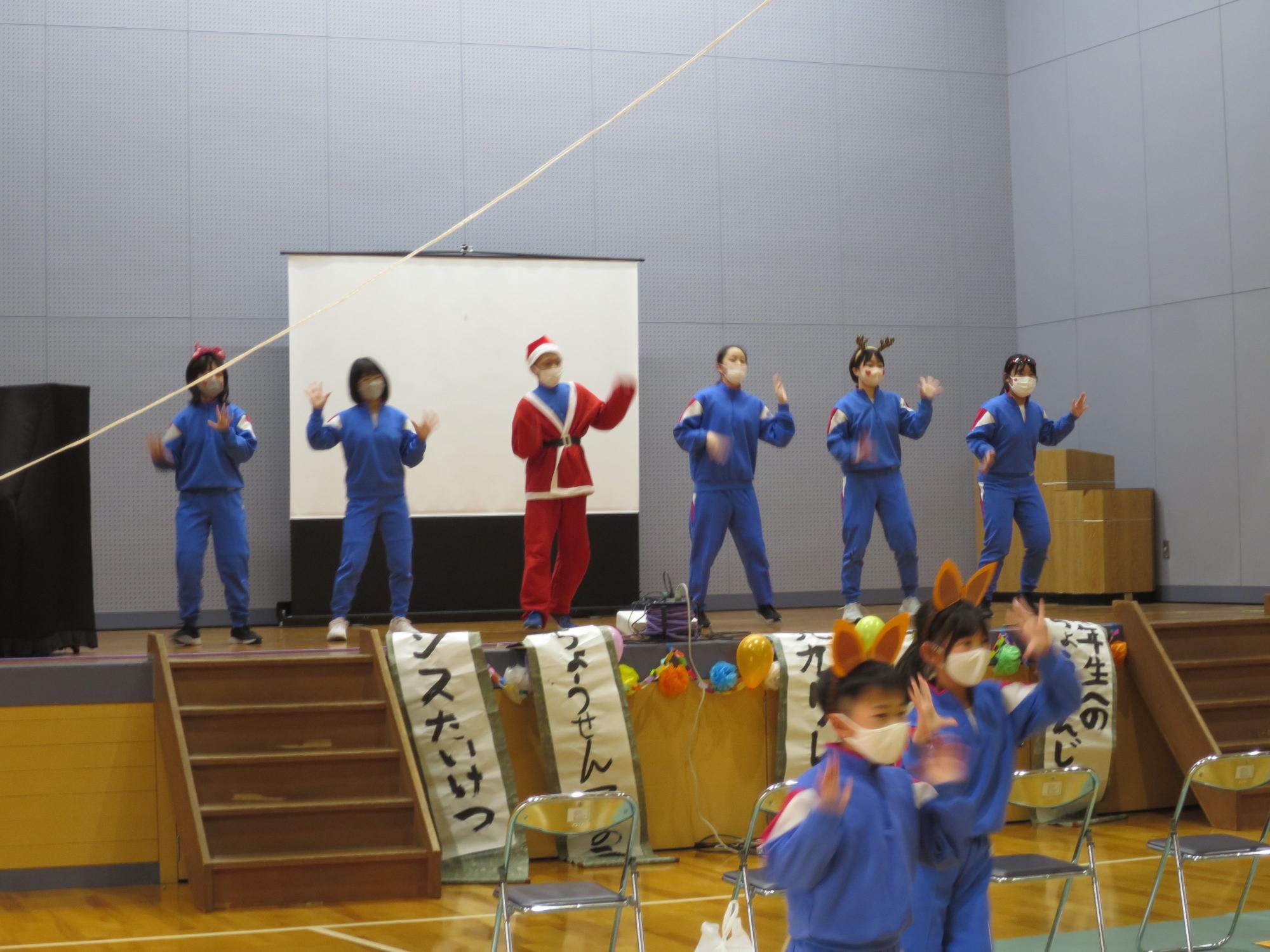 体育館の舞台で児童たちがサンタやトナカイの格好でダンスをしている写真