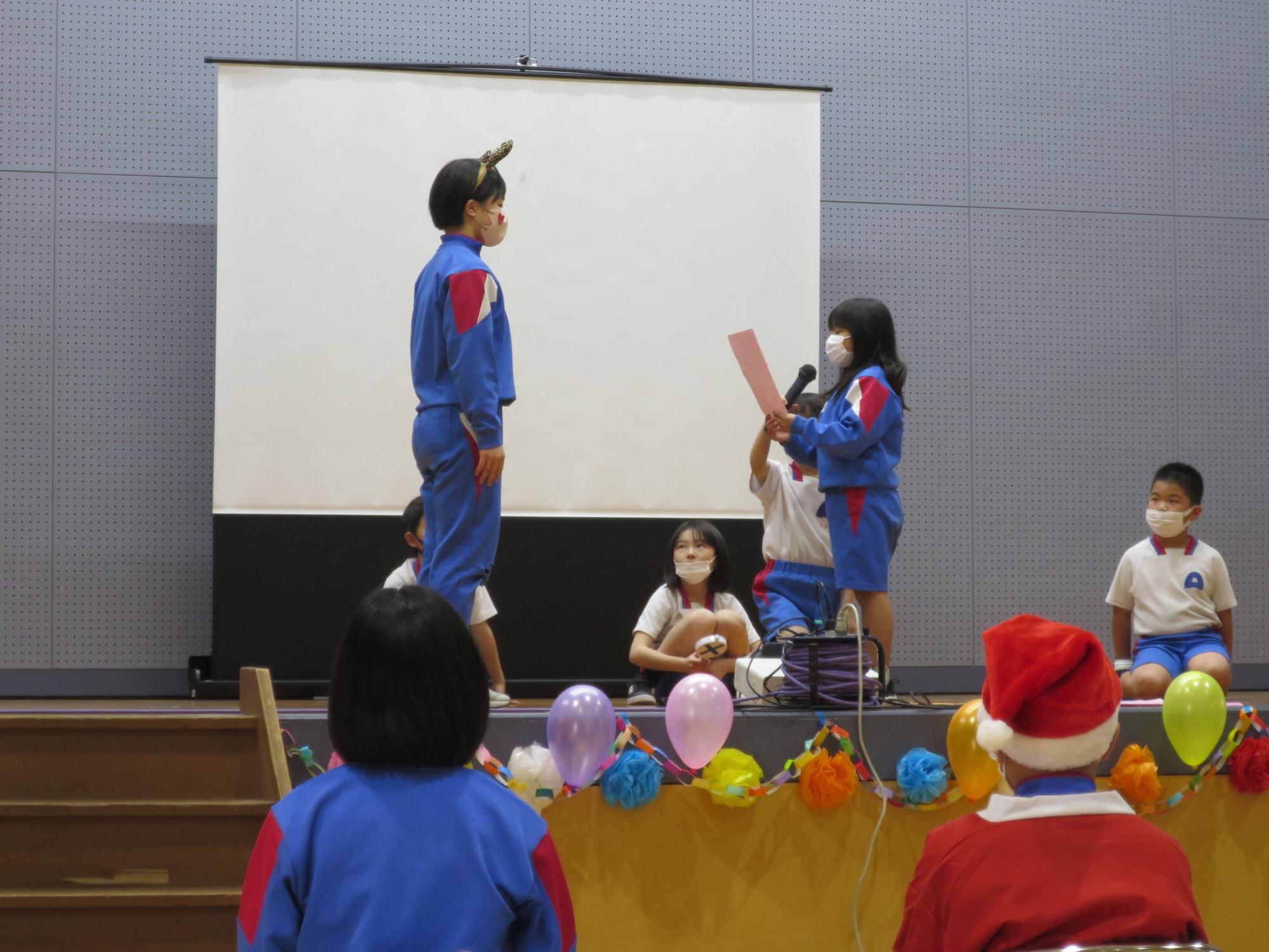 体育館の舞台で児童たちがマイクを使って発表している写真