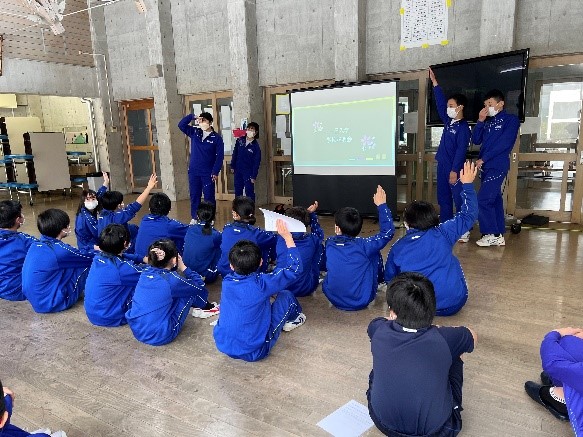 室内でモニターを使い手を挙げて発表を行う児童とそれを座って聞いている児童たちの写真