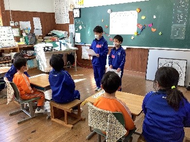 教室の中で青いジャージを着た二人の児童が発表をし、その様子を着席して聞いている複数の児童たちの写真