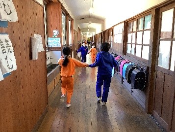 オレンジ色のジャージを着た児童と青色のジャージを着た二人の児童が手をつなぎ廊下を歩いている後ろ姿の写真
