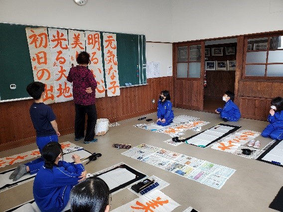 黒板にオレンジ色の字で様々な文字が書かれた書初めが貼られ、その前で教師が説明をし、その周りで話を聞く複数の児童たちの写真