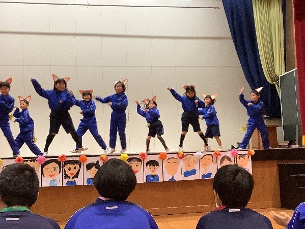 体育館の舞台上できつねの耳を着用し踊る青いジャージを着た複数人の児童たちの写真